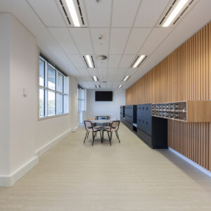 suspended grid section in a plasterboard ceiling. corridor with lockers, table and black and beige chairs; timber battens along right hand wall.
